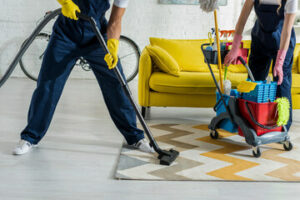 due addetti alla pulizia di un tappeto bianco giallo e grigio con divisa blu e guanti gialli passano un aspiratore 