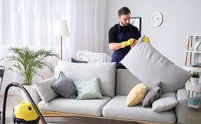 addetto alle pulizie con divisa blu e guanti gialli pulisce un divano grigio in un salone con una vaporella